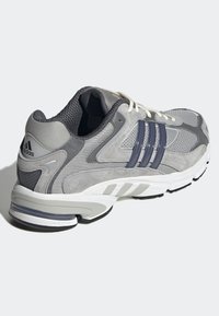Grey athletic shoe with mesh and suede materials, featuring three dark stripes, white laces, and a cushioned sole in contrasting colors.