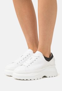 Vita klumpiga sneakers med en texturerad läderöverdel, svart hälaccent och tjock gummisula. Har runda snören och logodetaljer.