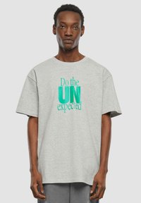 Grå bomull t-skjorte med korte ermer, med grafikk av "Do the UN expected" i teal og svart. Avslappet passform, rund hals.