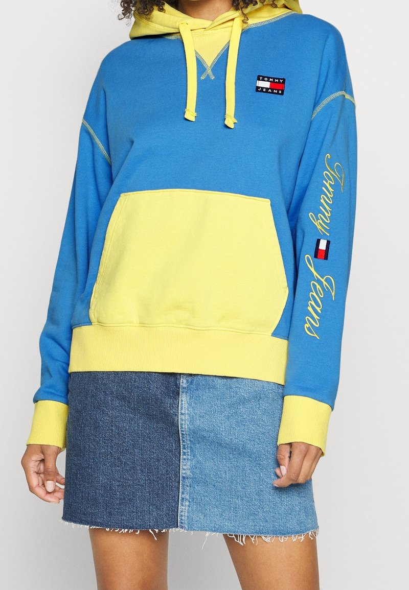 Sweat à capuche Tommy Jeans bleu et jaune avec poche avant et logo sur la poitrine et la manche, porté avec une mini-jupe en denim bicolore.