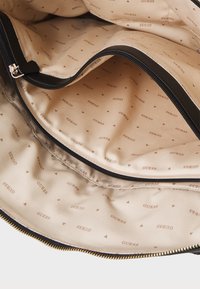 Zwarte ritstas met een beige binnenkant. De binnenkant bevat herhaalde bruine "GUESS" logo's en kleine driehoekige patronen. Glad, textuurstof.