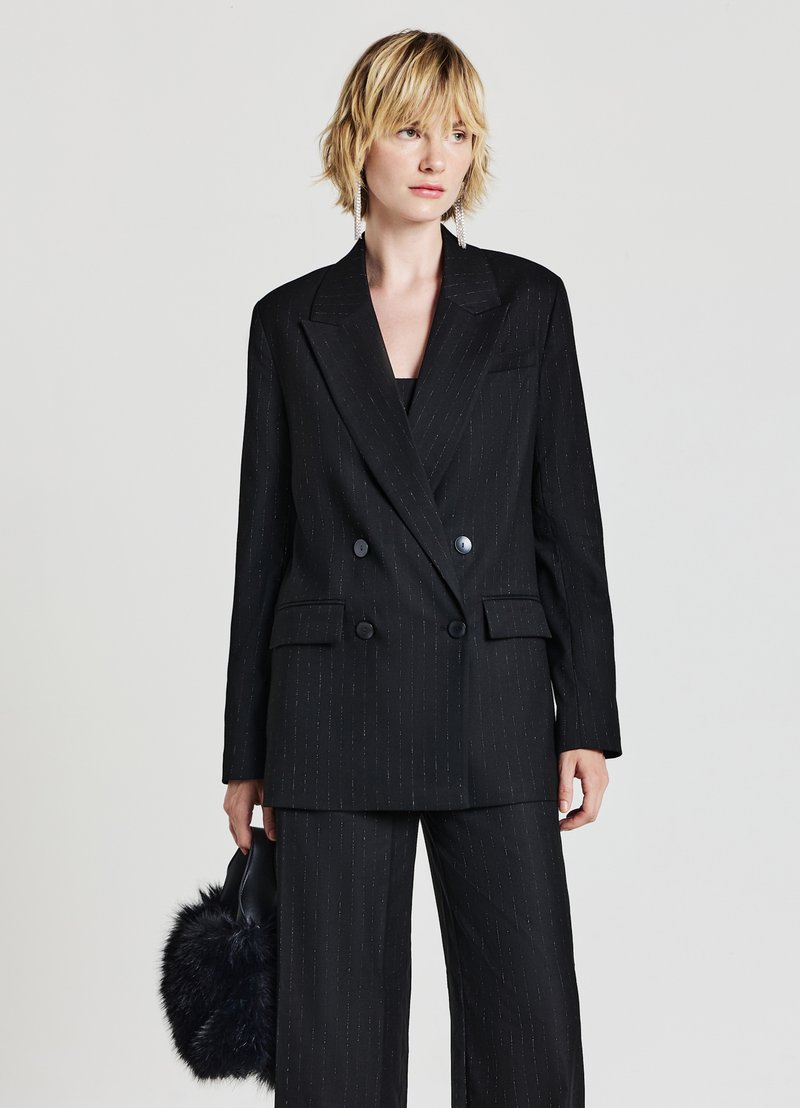 Completo a righe nere con giacca doppiopetto e pantaloni a gamba larga. Il materiale appare strutturato; il modello tiene una borsa in pelliccia sintetica nera.