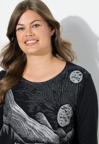 Hymyilevä nainen, jolla on laineikasta ruskeaa hiusta, pukeutunut mustaan paitaan, jossa on hopeisia abstrakteja maisemia ja pyöreitä kuvioita.