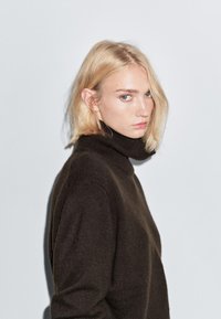 Ciemnobrązowy sweter z kołnierzem typu golf, o miękkiej fakturze, w luźnym kroju i wysokim kołnierzu. Modelka ma jasnoblond włosy i niebieskie oczy.