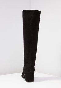 Des bottes en suédine noires au-dessus du genou présentent un talon carré, une texture lisse et une silhouette épurée agrémentée de détails de couture subtils.