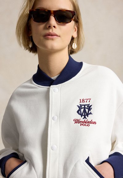 Young woman wearing tortoiseshell sunglasses, shell earrings, and a white Wimbledon Polo jacket with navy trim and embroidered logo.