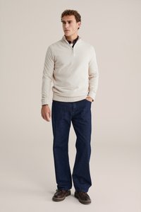 Helle beige Zip-Up-Strickjacke, dunkelblaue Jeans und braune Schuhe. Die Strickjacke weist ein strukturiertes Muster sowie gerippte Bündchen und Saum auf.