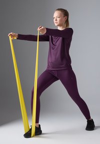 Camiseta atlética de manga larga morada y leggings ajustados; la modelo hace ejercicio con una banda de resistencia amarilla; zapatos negros sobre un fondo gris.