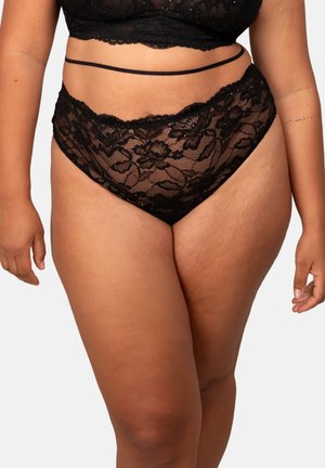 Curvy person wearing black floral lace high-waist panties and matching bralette with thin strap detail on waist, against a plain background.