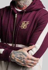 Hoodie zippé bordeaux et crème avec capuche, ornée d'un logo brodé en or sur la poitrine et de rayures contrastantes sur les manches. Mélange de coton.