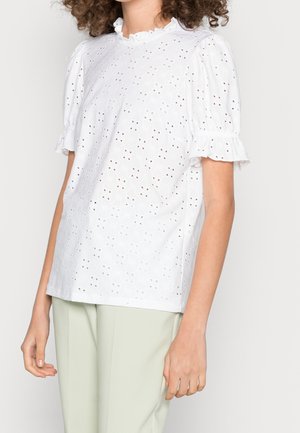 Blusa bianca in cotone con maniche corte a sbuffo, colletto arricciato e motivo ad ricamo ad occhiello. Indossata con pantaloni verde chiaro.