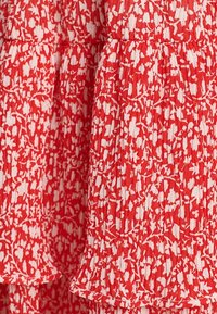 Punainen kukkakuosisempi kangas, jossa on valkoisia kukkia, pleatattu texture ja kerroksellinen helma. Kevyt, ideaalinen lämpimille säille.