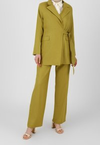 Olivgrüner maßgeschneiderter Blazer mit gebundenem Gürtel, kombiniert mit passenden weit geschnittenen Hosen. Glattes Material und strukturiertes Design. Sandalen mit quadratischen Absätzen.