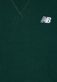 Zöld pulóver V-nyakú dizájnnal, bordás textúrával és kis, fehér hímezett "NB" logóval a bal oldalon. Puha, kötött anyag.