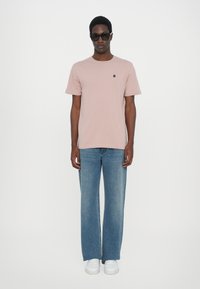 Kurzärmliges, hellrosa Baumwoll-T-Shirt mit kleinem gesticktem Logo auf der Brust, kombiniert mit weit geschnittenen blauen Jeans und weißen Sneakern.