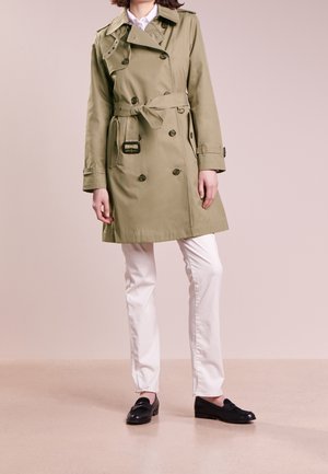 Person iført en khakifarvet trenchcoat med bælte, hvide bukser og sorte loafers, stående foran en ensfarvet beige baggrund.