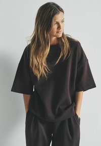 Tumši brūns pārāk liels T-krekls ar īsām piedurknēm un noapaļotu kakla izgriezumu, apvienots ar brīvā stila biksēm, kas izgatavotas no mīkstas auduma.