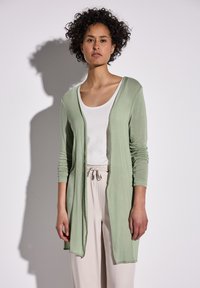 Lichtgroene lange cardigan met lange mouwen, open voorkant en geribbelde structuur, gecombineerd met een witte top en beige broek.