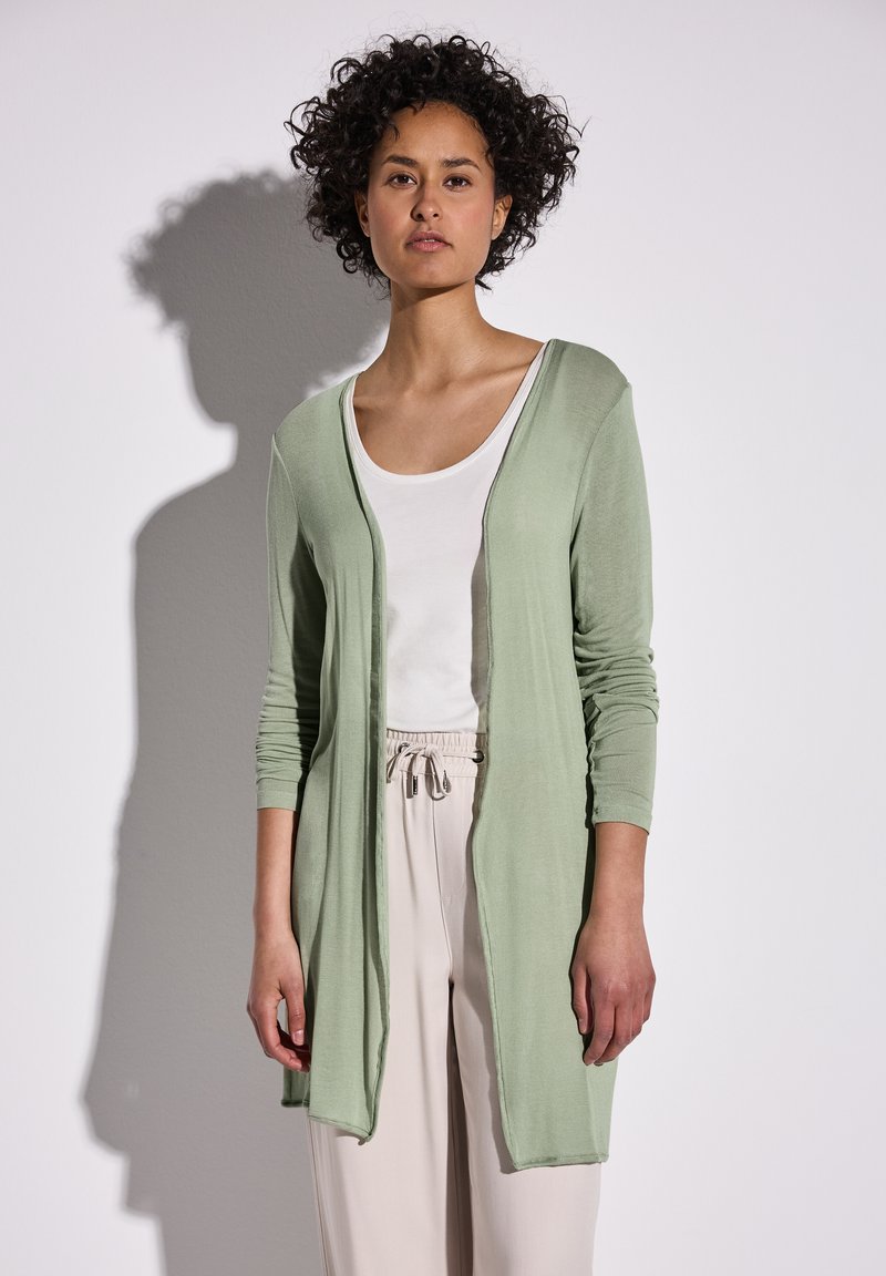 Lichtgroene lange cardigan met lange mouwen, open voorkant en geribbelde structuur, gecombineerd met een witte top en beige broek.