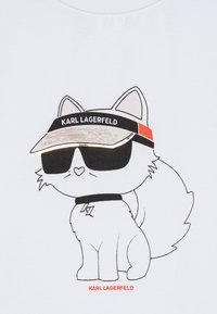 Bílý tričko s potiskem kreslené kočky v kšiltovce a slunečních brýlích, s nápisem "KARL LAGERFELD." Kočka má huňatý ocas a jednoduché rysy obličeje.