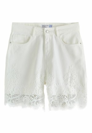 Kvinde iført hvide shorts med blonder, sort ærmeløs top og brune sandaler med åben tå og hæl står foran en lys grøn baggrund.