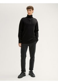 Schwarzer Strickpullover mit hohem Kragen und halbem Reißverschluss, ausgestattet mit einem strukturierten Vorderteil, kombiniert mit schwarzen trousers und Sneakers.