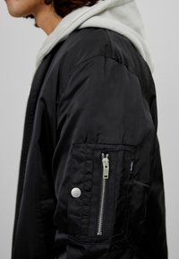 Svart bombarjacka med en slät, glänsande yta. Har en ficka med dragkedja på ärmen och en grå huva synlig under.