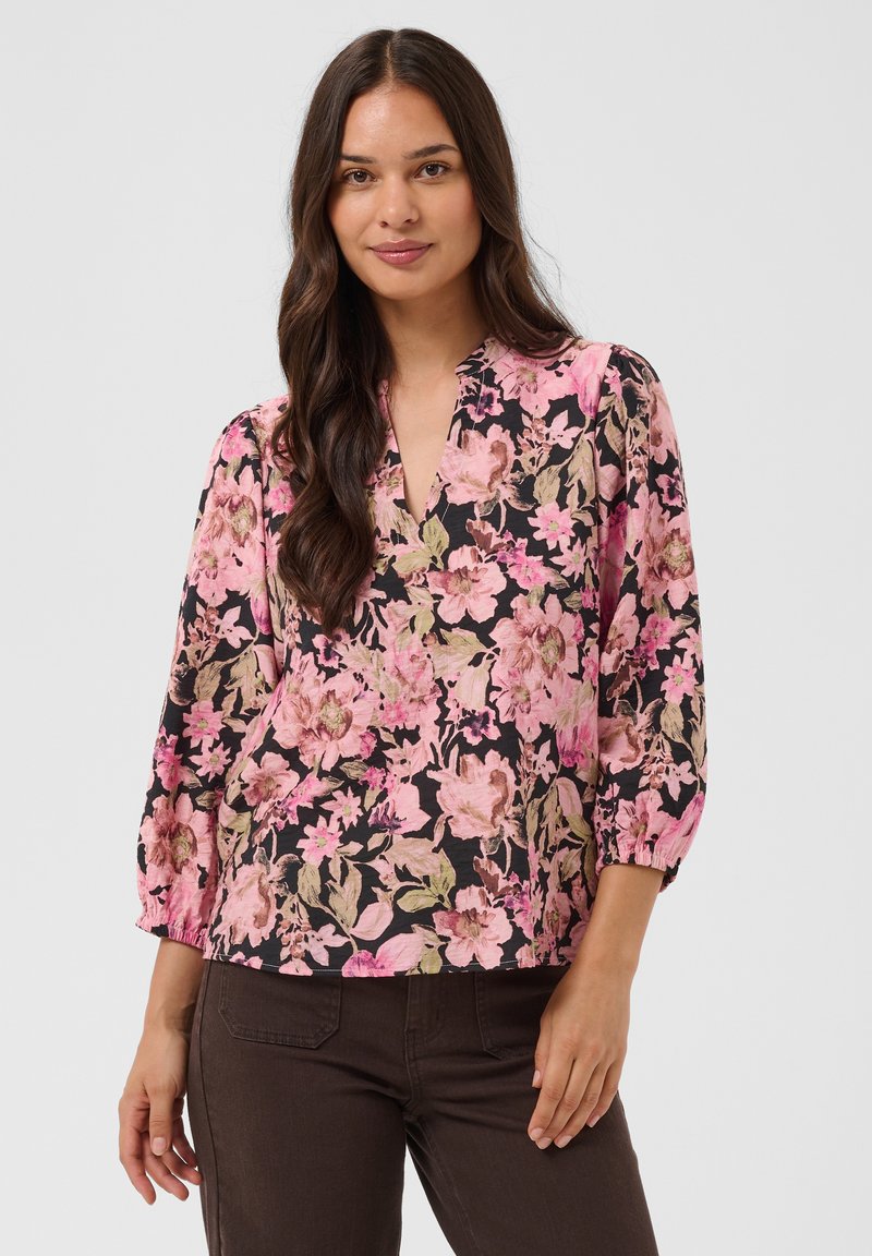 Femme aux longs cheveux foncés portant un chemisier floral rose et noir à manches trois-quarts et un pantalon marron foncé, debout devant un fond clair.