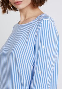 Kobieta ubrana w niebieską i białą poziomo prążkowaną koszulę z długim rękawem, z białymi guzikami na rękawach, na jednolitym tle.