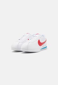White sneakers with red accents, featuring a textured surface, rounded toe, lace closure, and a blue stripe along the sole.