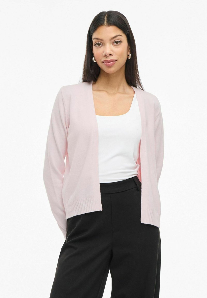Cardigan rosa claro con un diseño de cuello en V, puños acanalados y una textura suave, superpuesto a una blusa blanca y combinado con pantalones negros.