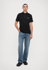 Svart polo skjorte med en tre-knapps placket og minimal merkevare, kombinert med lyseblå jeans og svarte sko. Enkel, uformell design.