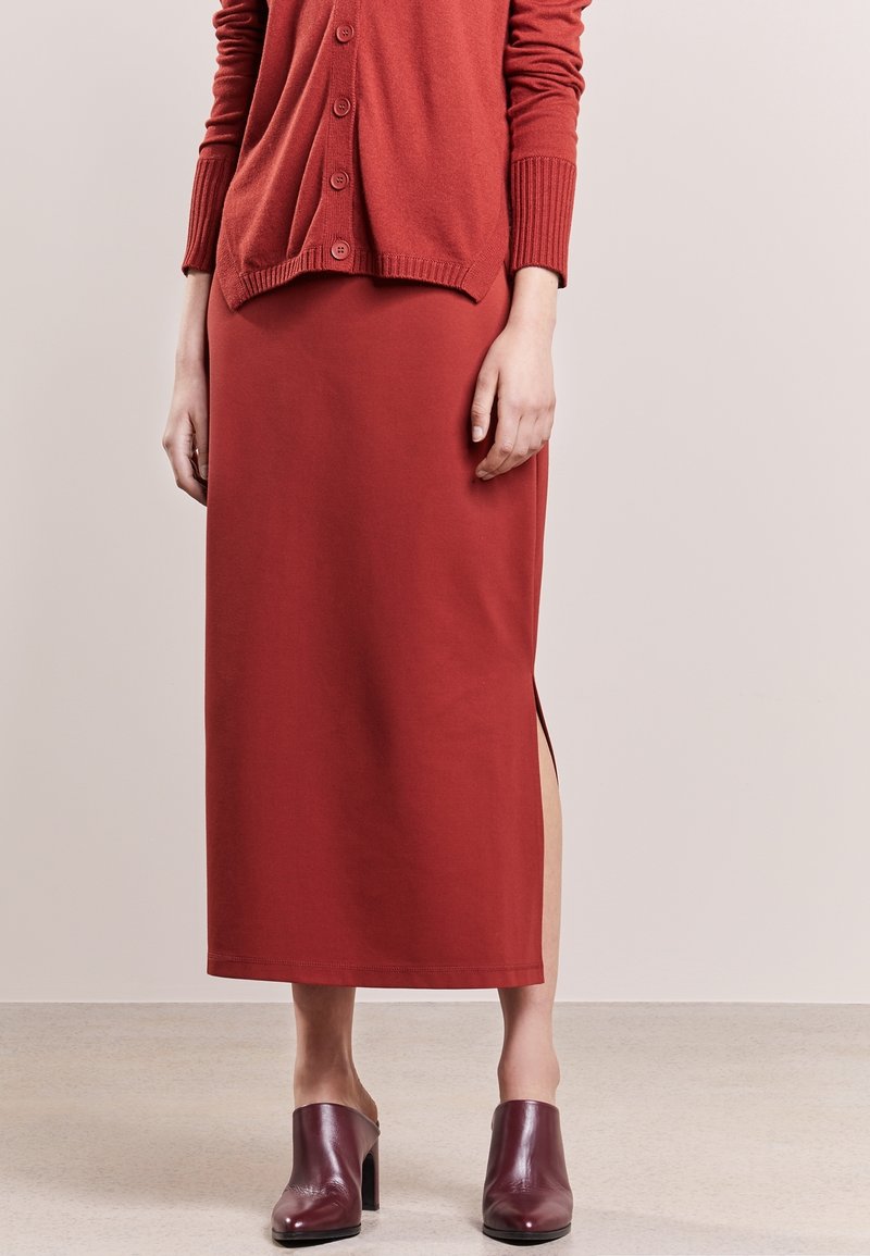 Femme portant une longue jupe couleur rouille avec une fente latérale, un cardigan boutonné assorti et des mules à talons rouge foncé sur un fond uni.