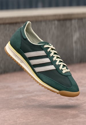 Green suede sneakers with mesh panels, three white stripes, cream laces, and a gum sole, featuring a rounded toe and low-profile design.