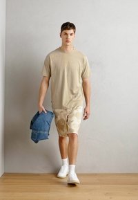 T-shirt beige, shorts à motif camouflage, baskets blanches et une veste en denim à la main ; sur un fond neutre avec un sol en bois.