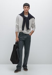 Gray sweater, navy scarf tied at the neck, dark denim jeans, and brown suede shoes. Holding a black suede tote bag. Minimal background.