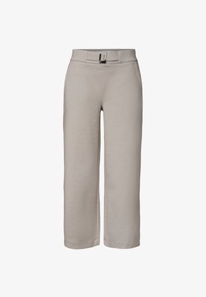 Person iført lysegrå skræddersyede bukser og en mørkerød top, der står mod en neutral baggrund med deres højre arm synlig.