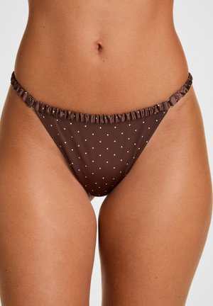 Brauner Bikinihöschen mit weißen Punkten und gerafftem Bund, getragen an den Hüften eines Models vor einfachem Hintergrund.