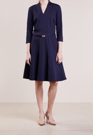 Kobieta w granatowej sukience do kolan z rękawami do łokcia i wąskim czarnym paskiem, w połączeniu z cielistymi szpilkami.