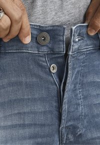 Ljusblå denimjeans med knappstängning och metallbeslag. Tyget har en sliten textur med synliga sömmar och fickor.