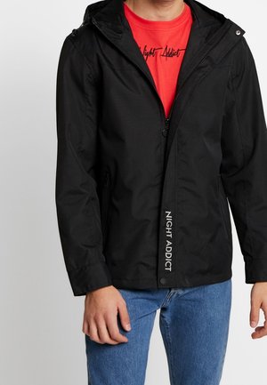 Mężczyzna w czarnej kurtce z kapturem z napisem "NIGHT ADDICT" i czerwonej koszulce z czarnym nadrukiem "Night Addict", w połączeniu z niebieskimi dżinsami.