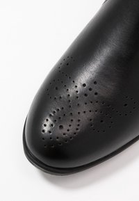 Scarpa di pelle nera con una texture liscia e perforazioni decorative in un motivo a spirale sulla punta. Forma della punta arrotondata, suola piatta.