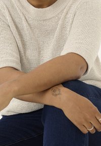 Vaaleanbeige neulepaita, jossa on leveä pääntie ja lyhyet hihat, puettuna tummien farkkujen päälle. Toisessa kädessä näkyy kukkakuvioinen tatuointi, korut ovat vähäiset.