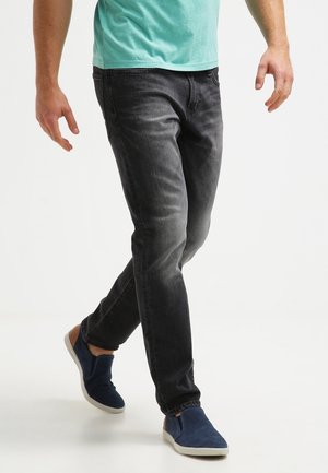 Henkilö kävelee kuluneissa mustissa farkuissa, teal-värisessä paidassa ja sinisissä liukusissa, joissa on valkoiset pohjat, yksinkertaisella valkoisella taustalla.