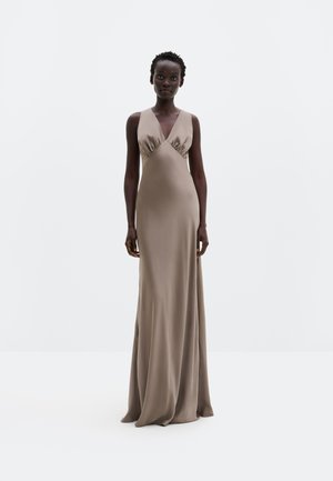 Frau mit kurzen lockigen Haaren, die ein ärmelloses, taupefarbenes Satinkleid mit tiefem V-Ausschnitt trägt und vor einem schlichten weißen Hintergrund steht.