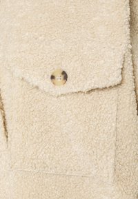 Beiger Kunstfellstoff mit strukturierter Oberfläche, ausgestattet mit einer großen Brusttasche und einem runden, hellen Schildpattknopf-Detail.