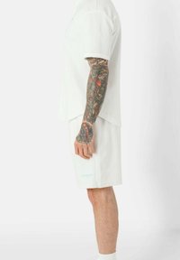 T-shirt et short de sport blancs, en tissu texturé, avec un logo sur l'ourlet gauche. Détails visibles des manches et des tatouages sur les bras.