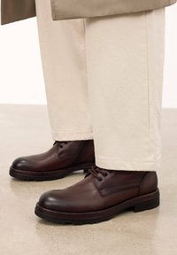 Pantofi din piele maro cu boturi rotunde, design cu sireturi și părți texturate. Purtați cu pantaloni de culoare crem, punând în evidență un stil casual.