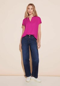 Top fucsia a maniche corte con scollatura a V, abbinato a jeans dritti blu scuro. Il modello indossa sneakers bianche, in piedi su uno sfondo chiaro.