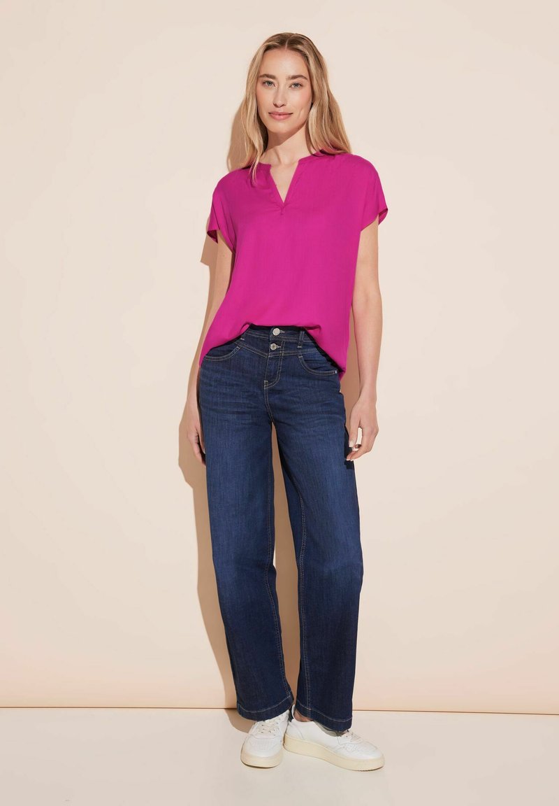 Top fucsia a maniche corte con scollatura a V, abbinato a jeans dritti blu scuro. Il modello indossa sneakers bianche, in piedi su uno sfondo chiaro.
